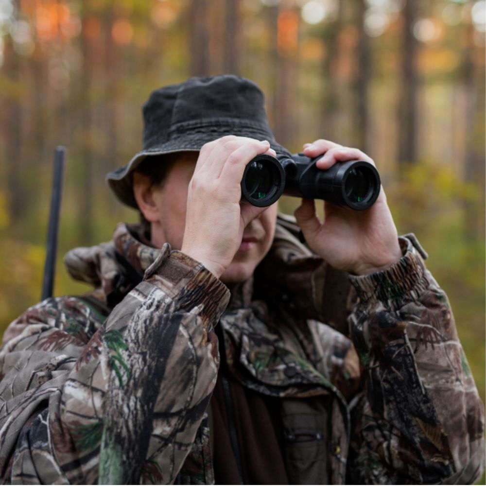 A person in camouflage clothing and a bucket hat looks through black binoculars in a forest setting with autumn-colored trees in the background.