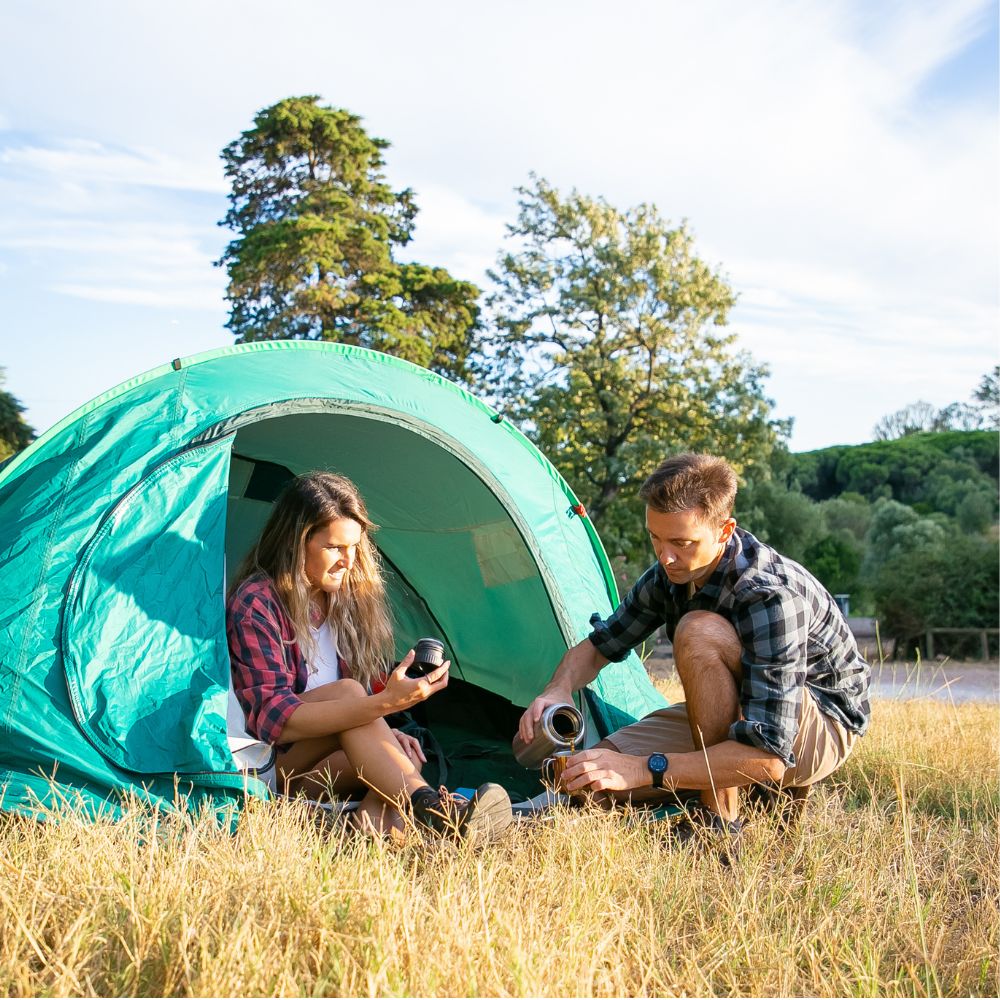 Two people sitting outside a green tent in a grassy field with trees in the background.