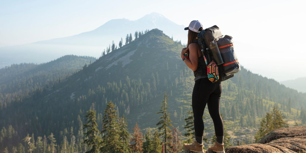 A hiker stands on a rocky mountain ridge wearing a large black and orange hiking backpack, looking out over a forested mountain range under a hazy sky.