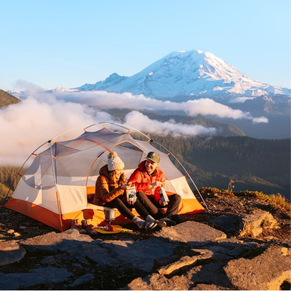 A dome camping tent is pitched on a rocky mountain summit with two people sitting inside it, facing a snow-capped mountain and clouds in the background.
