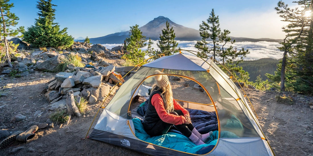 A woman sits inside a dome camping tent set up outdoors on a rocky mountain slope, with evergreen trees, a mountain peak, and a sea of clouds in the background at sunrise.