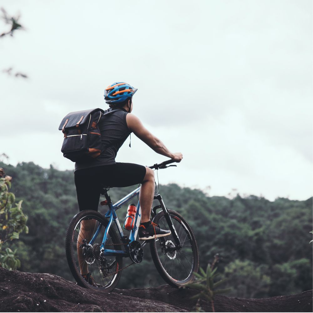 A person riding a blue mountain bike on a rocky trail, viewed from behind, wearing a black outfit, a blue and orange helmet, and a black backpack, with a forest in the background under an overcast sky.