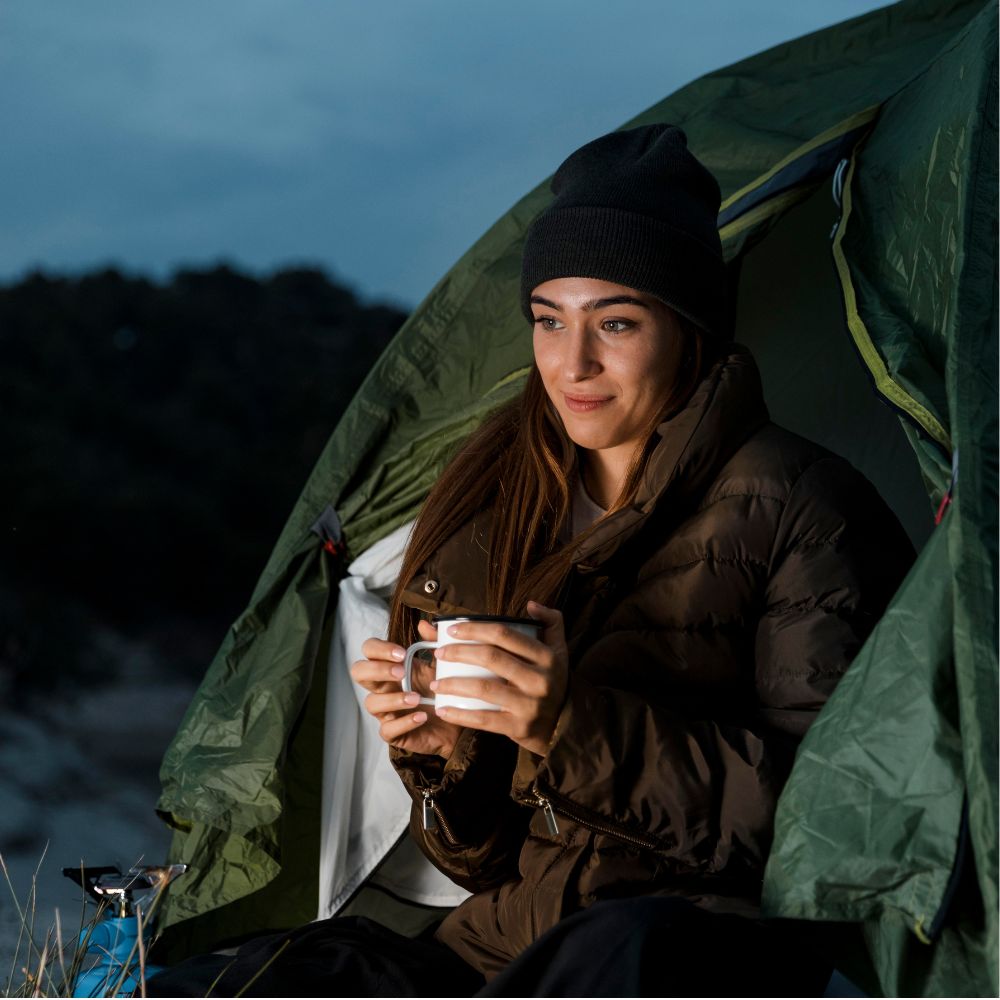 A woman wearing a black beanie and brown puffer jacket sits inside an olive green camping tent, holding a white mug in a dim outdoor setting at dusk.