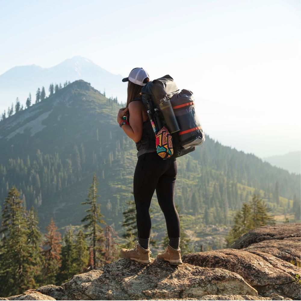 A woman stands on a rocky mountain peak wearing a large hiking backpack, black leggings, and brown boots, with forested hills and distant mountains in the background.