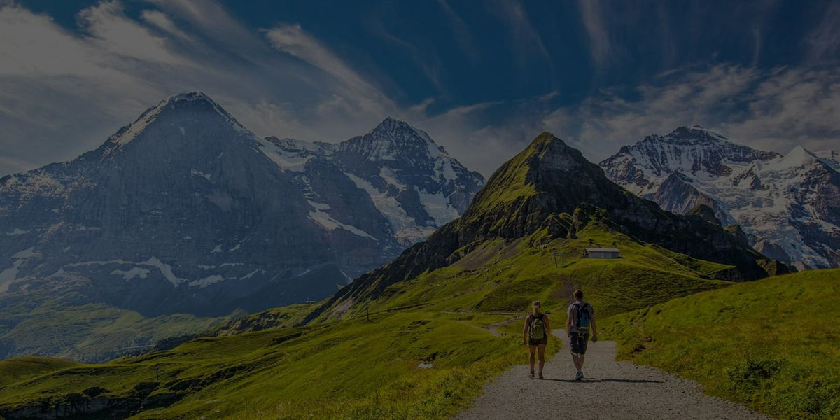 A scenic mountain landscape with snow-capped peaks, a green alpine meadow, and a path where two small hikers are walking. The image has a dark overlay, likely for a digital background or graphic asset.